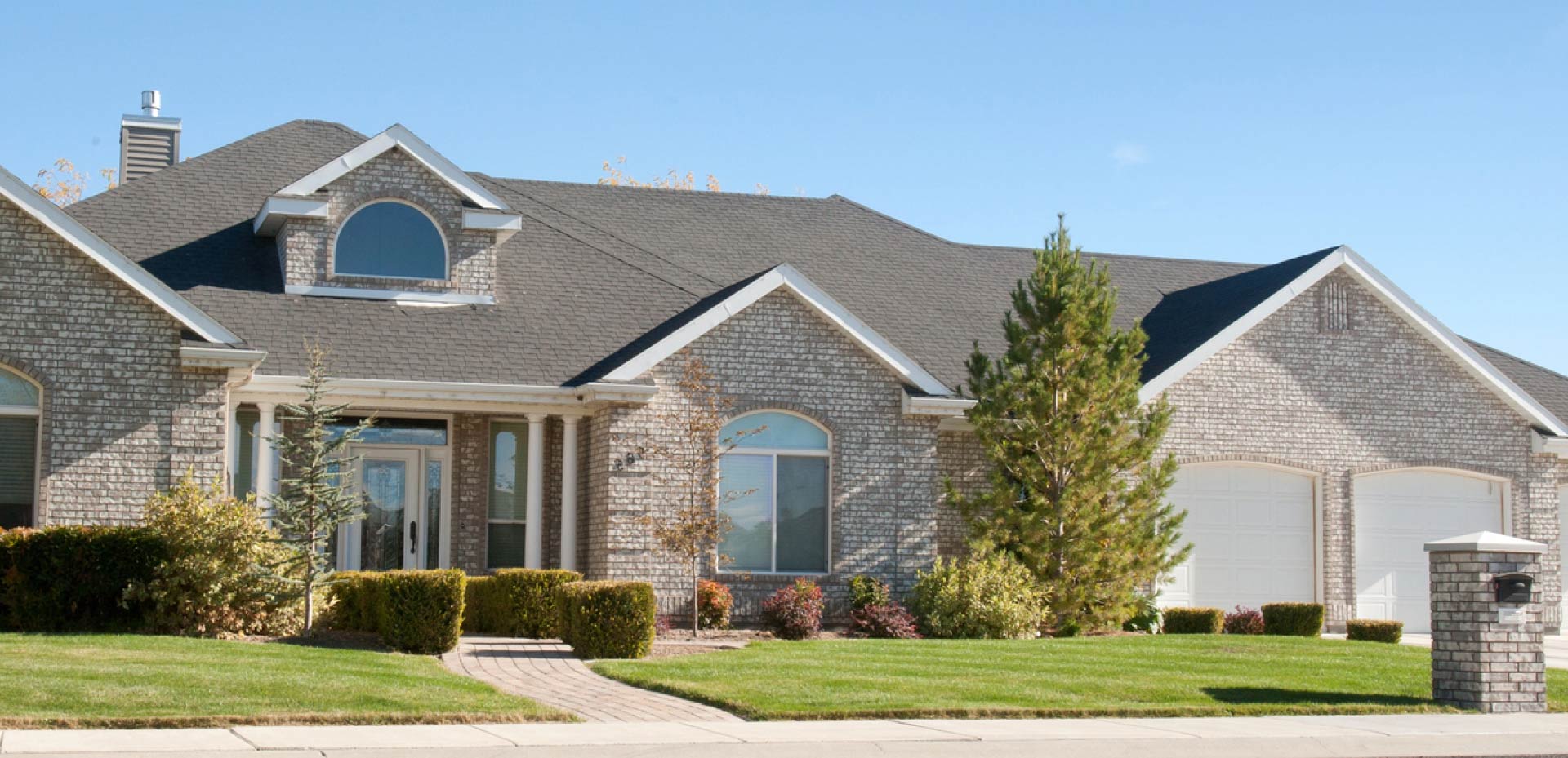 Large single-story suburban home featuring a pristine dark grey architectural shingle roof system on a brick exterior with manicured landscaping under a blue sky.