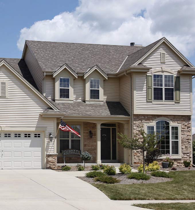 Two-story suburban home featuring a durable grey architectural shingle roof with dormers and stone facade accents under a cloudy blue sky.
