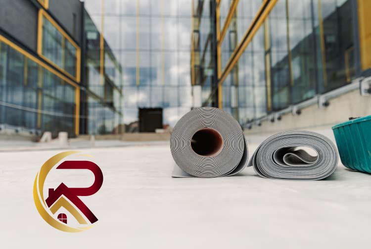Rolls of white and grey TPO roofing membrane and a green tool container sitting on a partially completed flat roof deck with a modern glass building facade in the background, featuring a red and gold company logo.