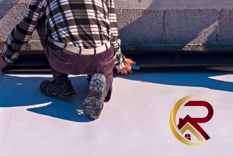 Professional roofer in work clothes kneeling while installing a black rubberized edge or membrane layer over a white TPO flat roof near a cinderblock wall, with a red and gold company logo.